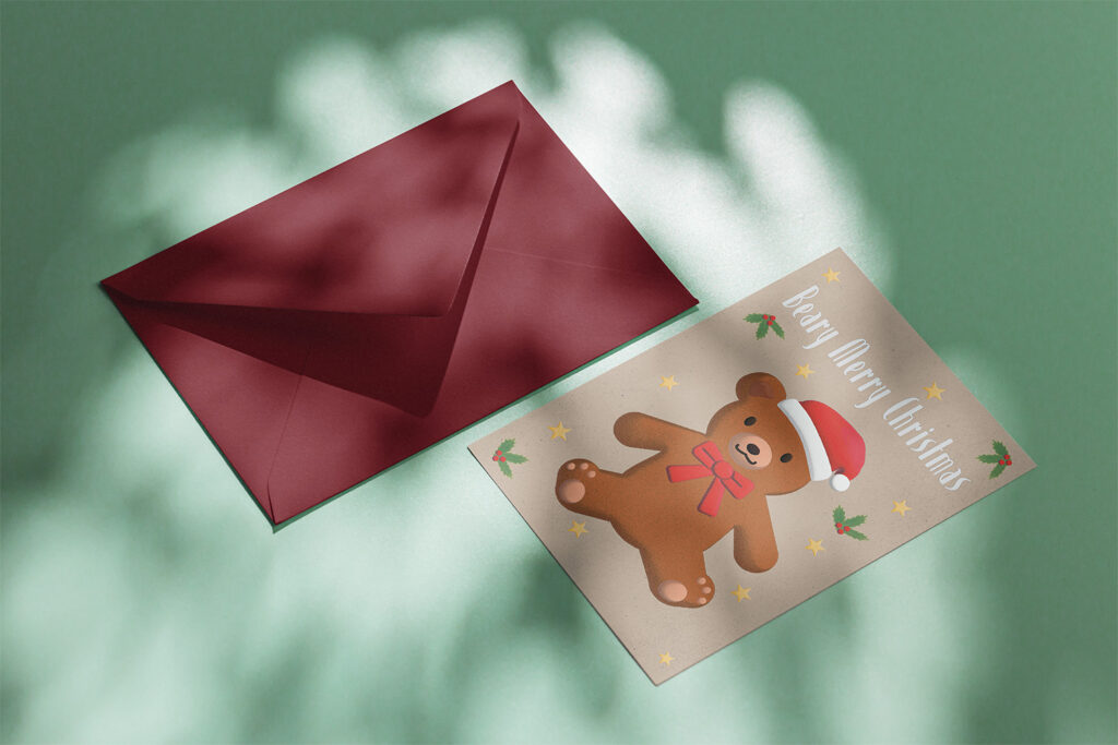 A Christmas greeting card featuring a cute teddy bear wearing a Santa hat and red bow, accompanied by festive holly and stars, next to a maroon envelope on a green surface.